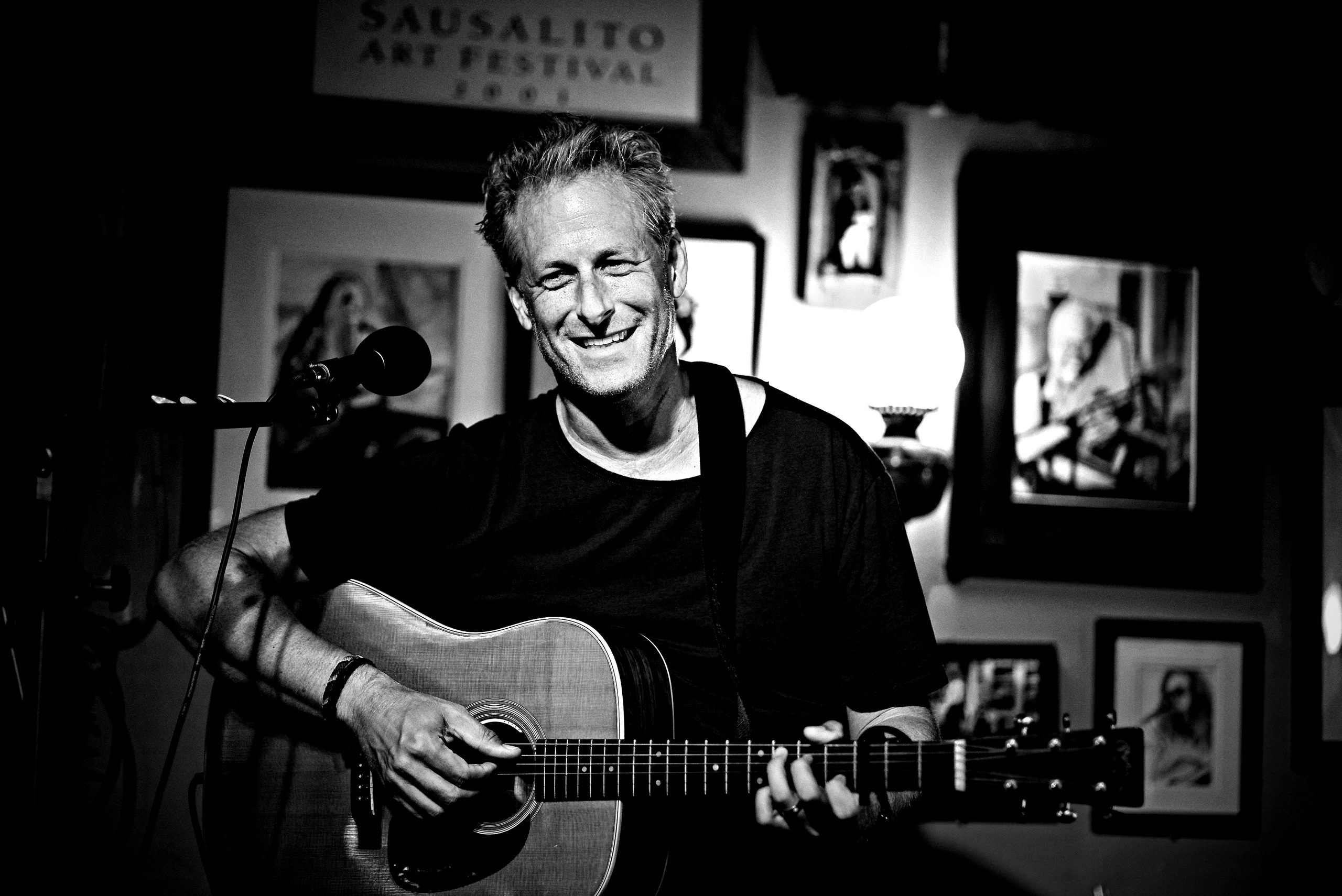 Pascal smiling with his acoustic guitar on stage at the Sausalito Art Festival, black and white